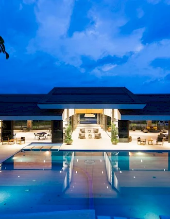 A luxurious house with a large, illuminated pool at night, surrounded by outdoor seating and a clear sky reflected on the water.
