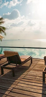 A serene tropical poolside with wooden deck chairs, umbrellas, palm trees, and a calm ocean view under a partly cloudy sky.