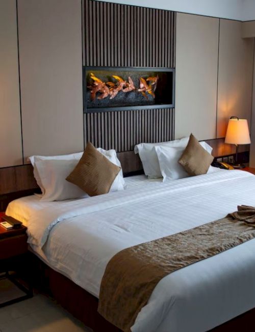 A hotel room with a large bed, two lamps on side tables, cushion accents, and a lit wall feature, with curtains over a window.