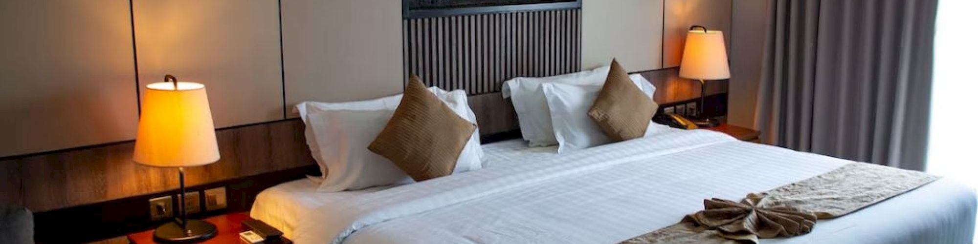A hotel room with a large bed, two bedside tables with lamps, and a window with curtains, featuring a small wall-mounted display.