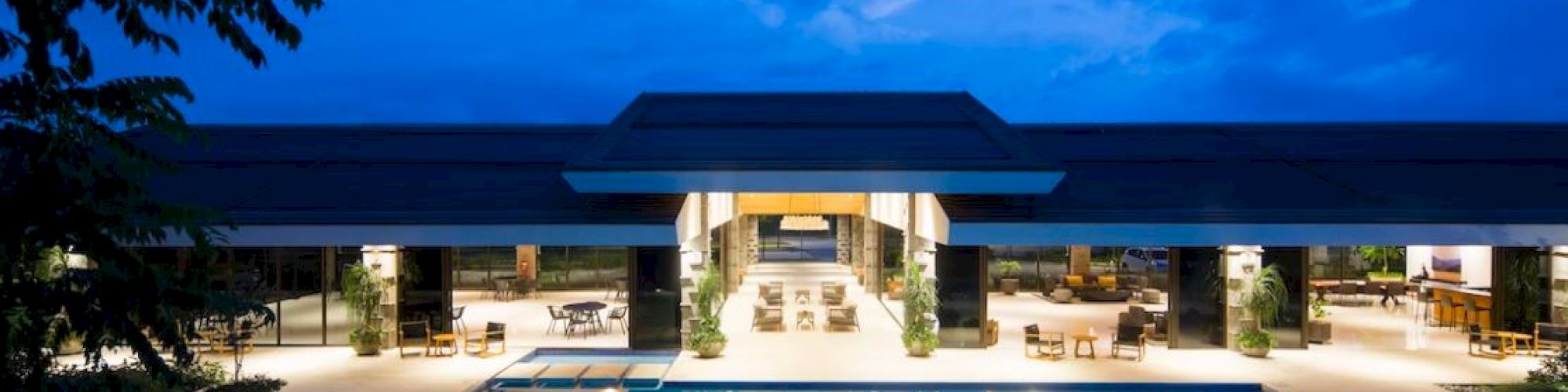 The image shows a modern building with large open areas, a lit swimming pool in front, and a darkening sky above.