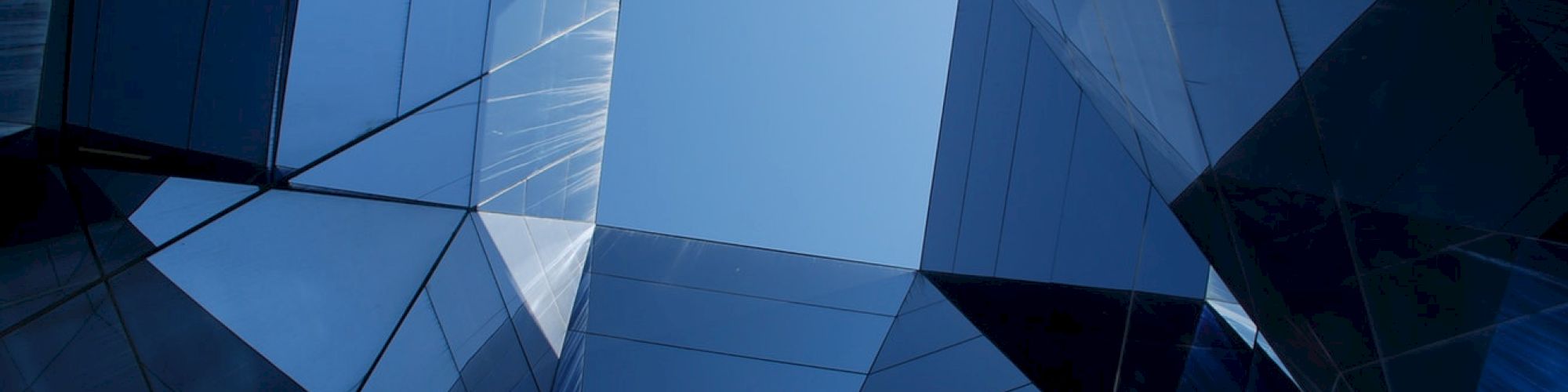 The image shows a geometric arrangement of reflective glass panels, forming a skylight that frames the blue sky above.