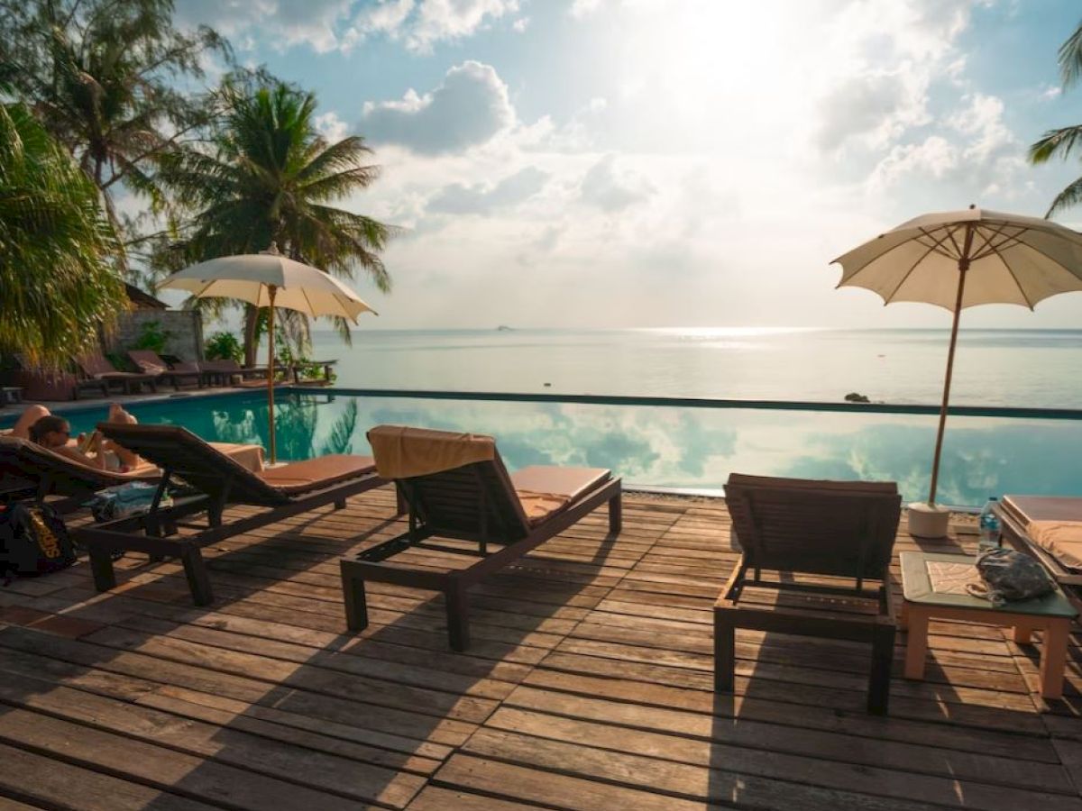 A serene beachside scene with lounge chairs, umbrellas, and a pool overlooking the ocean under a partly cloudy sky.