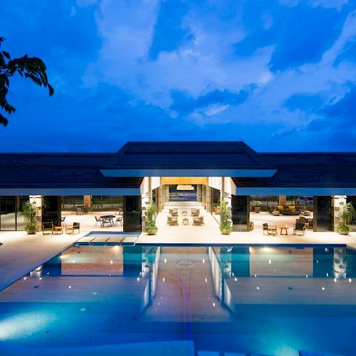 A luxurious outdoor pool area with loungers and a modern building is lit up against a twilight sky, surrounded by trees and open spaces.