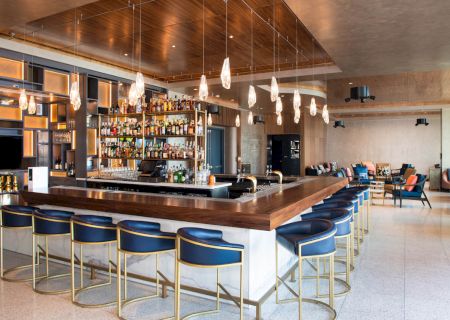 A modern bar with a wooden countertop, blue and gold barstools, pendant lights, and an extensive liquor selection.