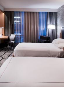 The image shows a modern hotel room with two double beds, a wall-mounted TV, a desk with a chair, and patterned curtains and wallpaper.