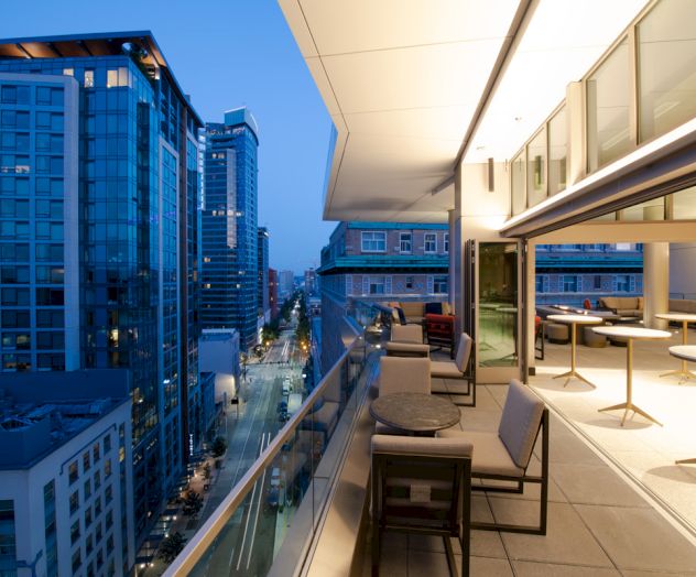 A modern cityscape view from a balcony at dusk, with tall buildings, illuminated streets, and a sleek outdoor seating area.