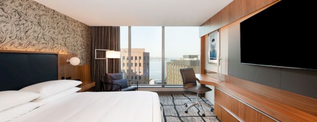 A modern hotel room features a large bed, desk, chair, TV, wall art, and a window offering a city and water view, with a stylish design.