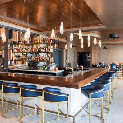 A modern bar with hanging lights, blue stools, and a wide variety of bottles on shelves. Cozy seating area in the background.