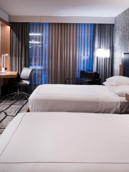 The image shows a modern hotel room with two beds, a TV, a desk with a chair, and stylish decor featuring patterned wallpaper.