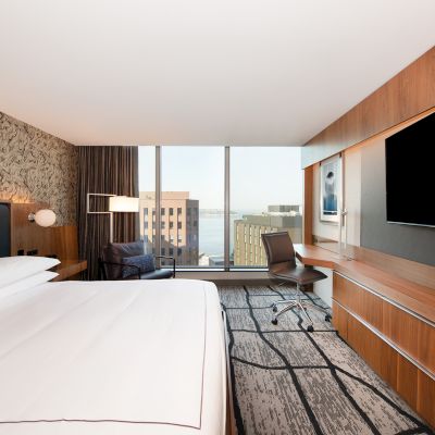 A modern hotel room with a large bed, a desk, a chair, a flat-screen TV, and a window with a city view completes the scene.