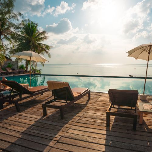 Poolside deck with lounge chairs and umbrellas facing a calm ocean under a sunny sky, surrounded by palm trees.