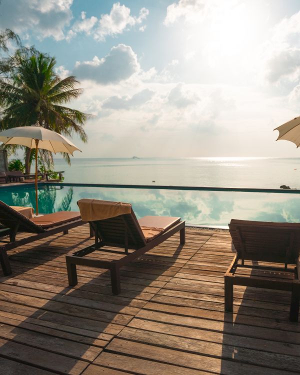 Poolside deck with lounge chairs and umbrellas facing a calm ocean under a sunny sky, surrounded by palm trees.