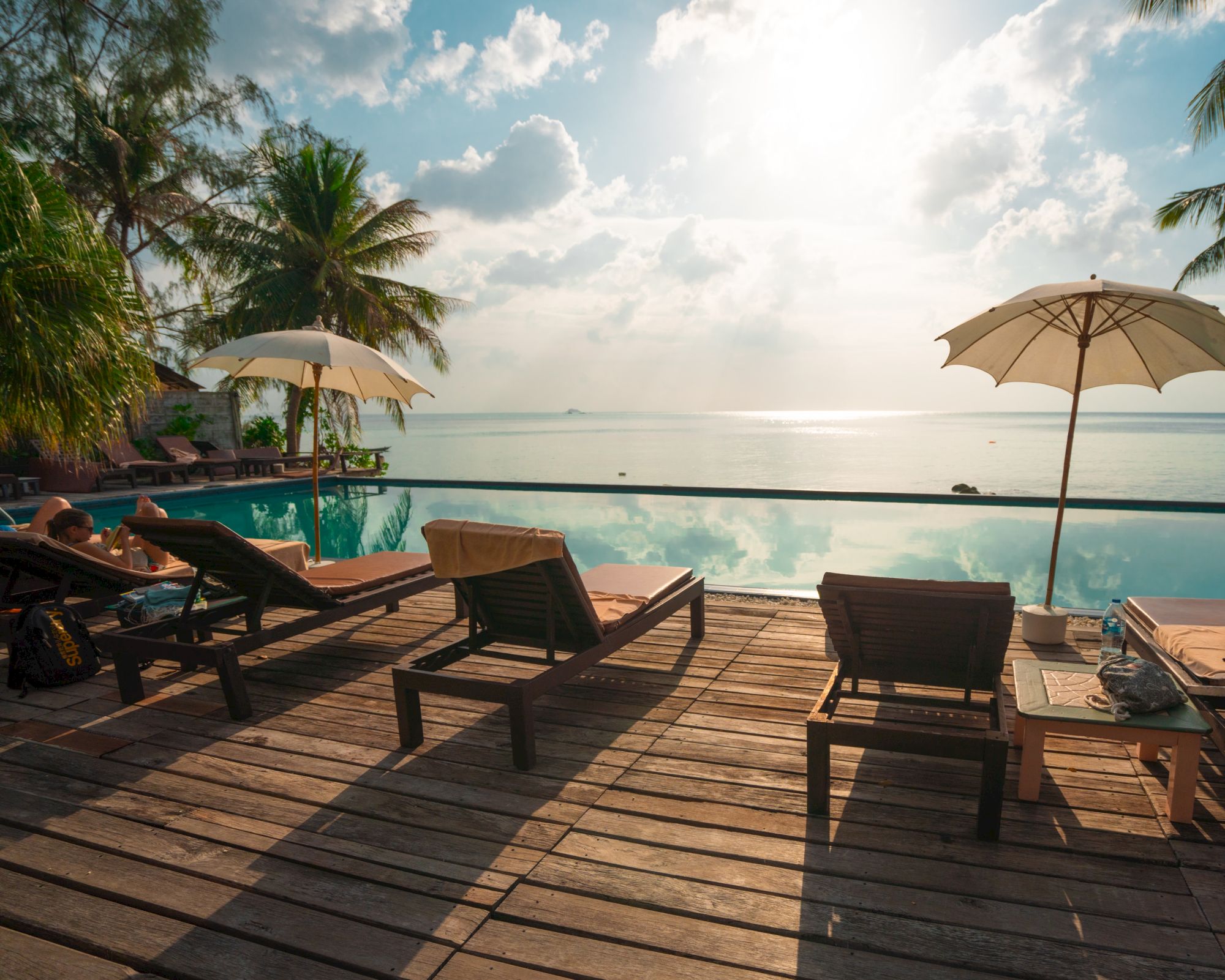 Sunlit poolside with lounge chairs, umbrellas, and palm trees overlooking a tranquil ocean under a partly cloudy sky.