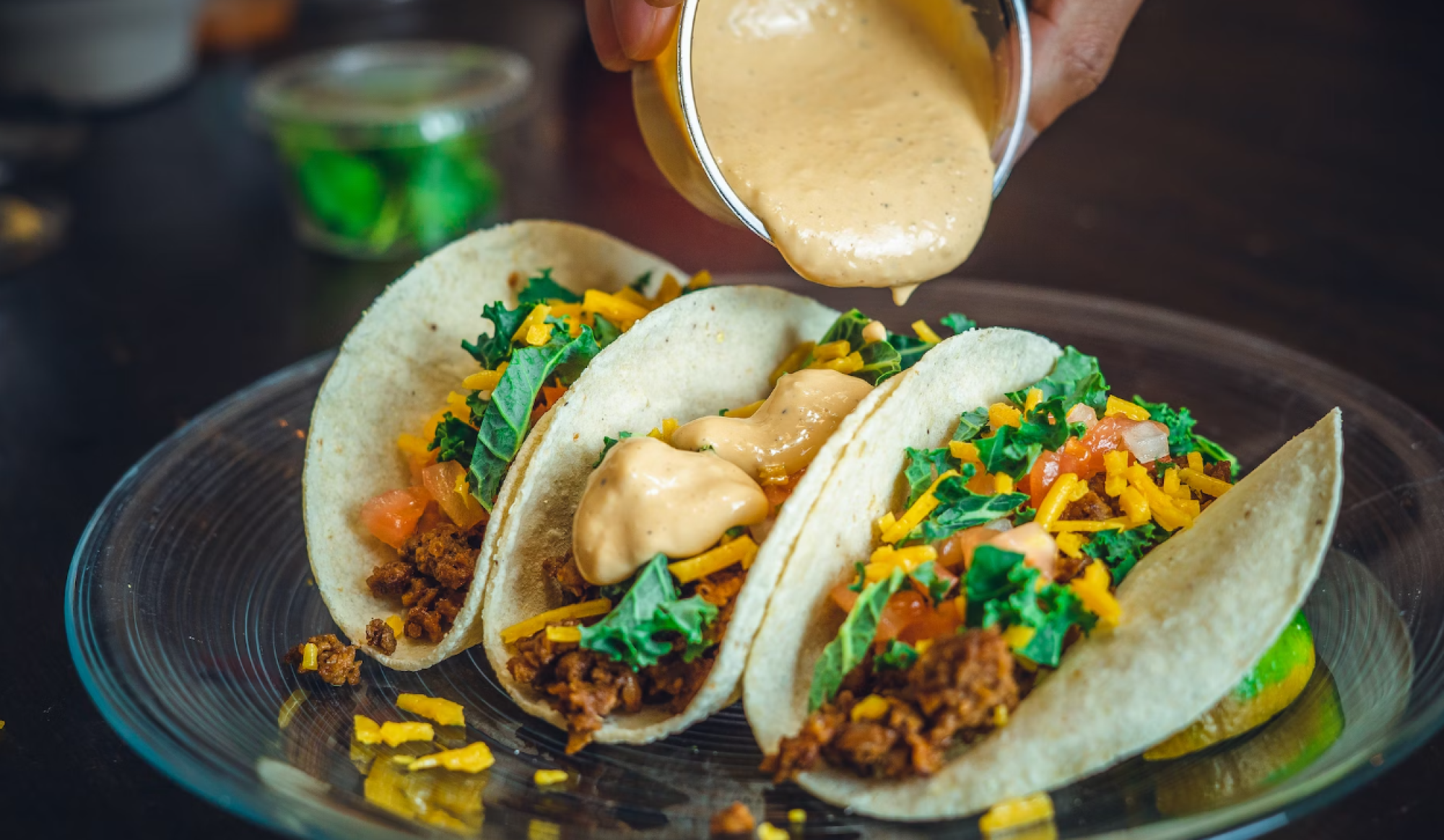 Three tacos filled with meat, lettuce, tomatoes, shredded cheese, and sauce being poured on top, displayed on a glass plate.
