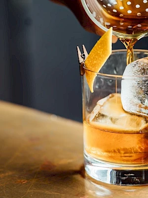 A cocktail is being poured into a glass with ice and a lemon twist garnish, using a strainer.