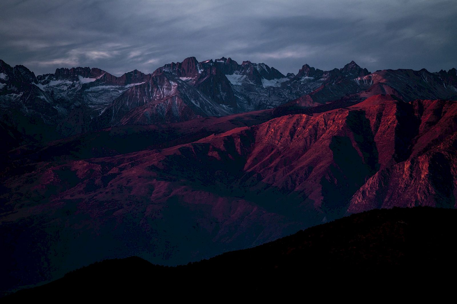 A mountain range under a dramatic sky, bathed in soft, reddish sunlight, with rugged peaks and shadows adding depth to the scene.