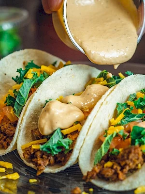 Three tacos filled with ground meat, cheese, lettuce, and tomatoes, topped with a creamy sauce being poured over them.