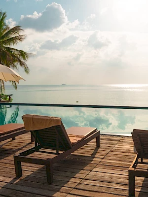 Poolside scene with lounge chairs, umbrellas, and palm trees overlooking a serene ocean view under a partly cloudy sky.
