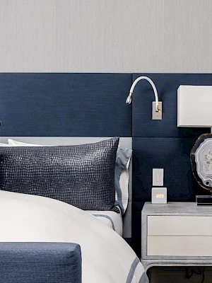 A modern bedroom features a blue headboard, white bed, dark pillows, a nightstand with a lamp, and a wall-mounted reading light.