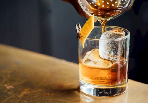 A cocktail being poured into a glass with large ice cubes and garnished with a twist of citrus peel on the rim.