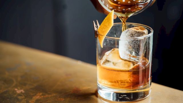 A cocktail being poured into a glass with large ice cubes and garnished with a twist of citrus peel on the rim.