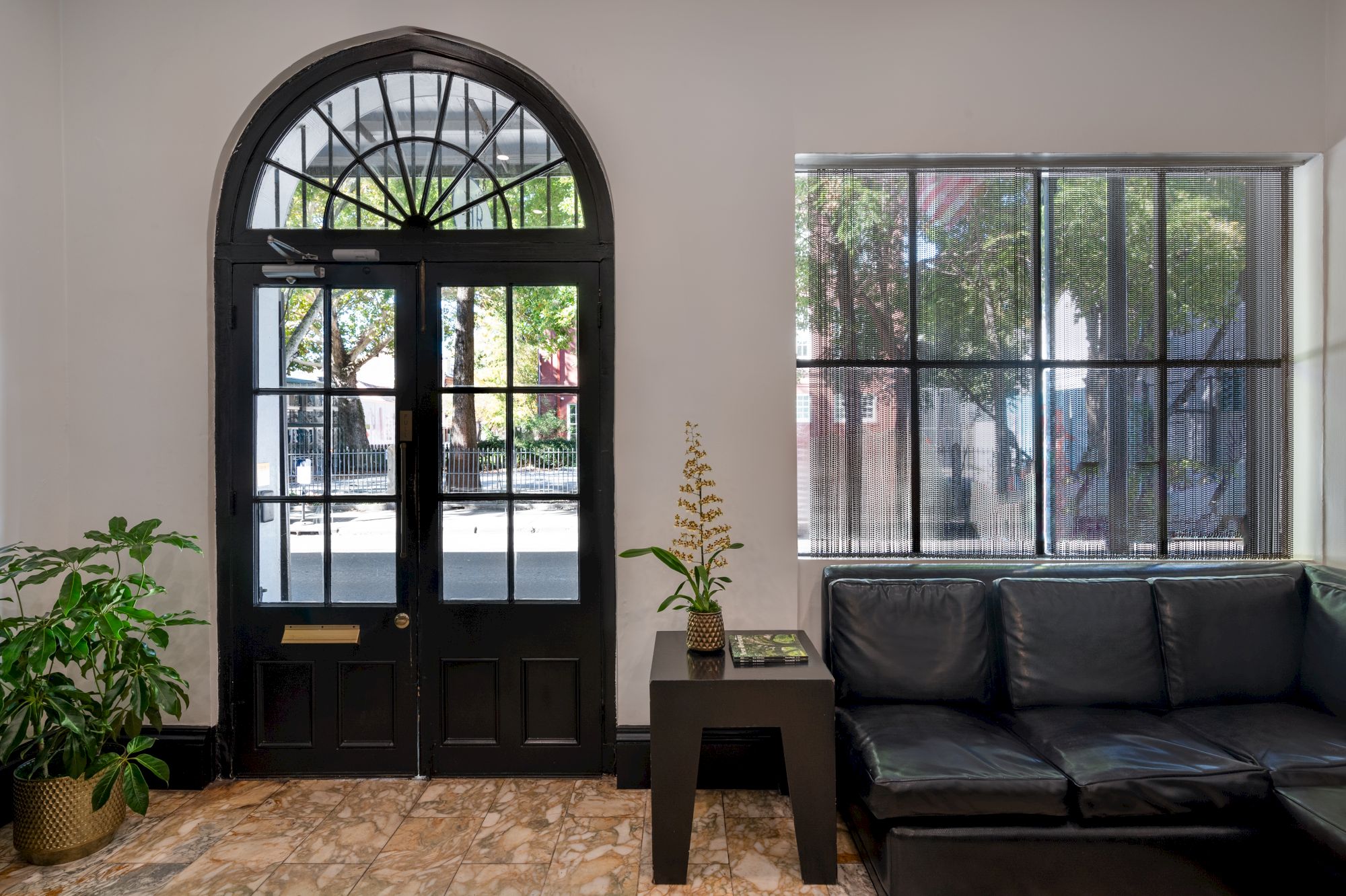 A black front door with glass panes, a large arched transom, a windowed wall to the right, a black leather sofa, and a small table with a potted plant in a bright, airy lobby.