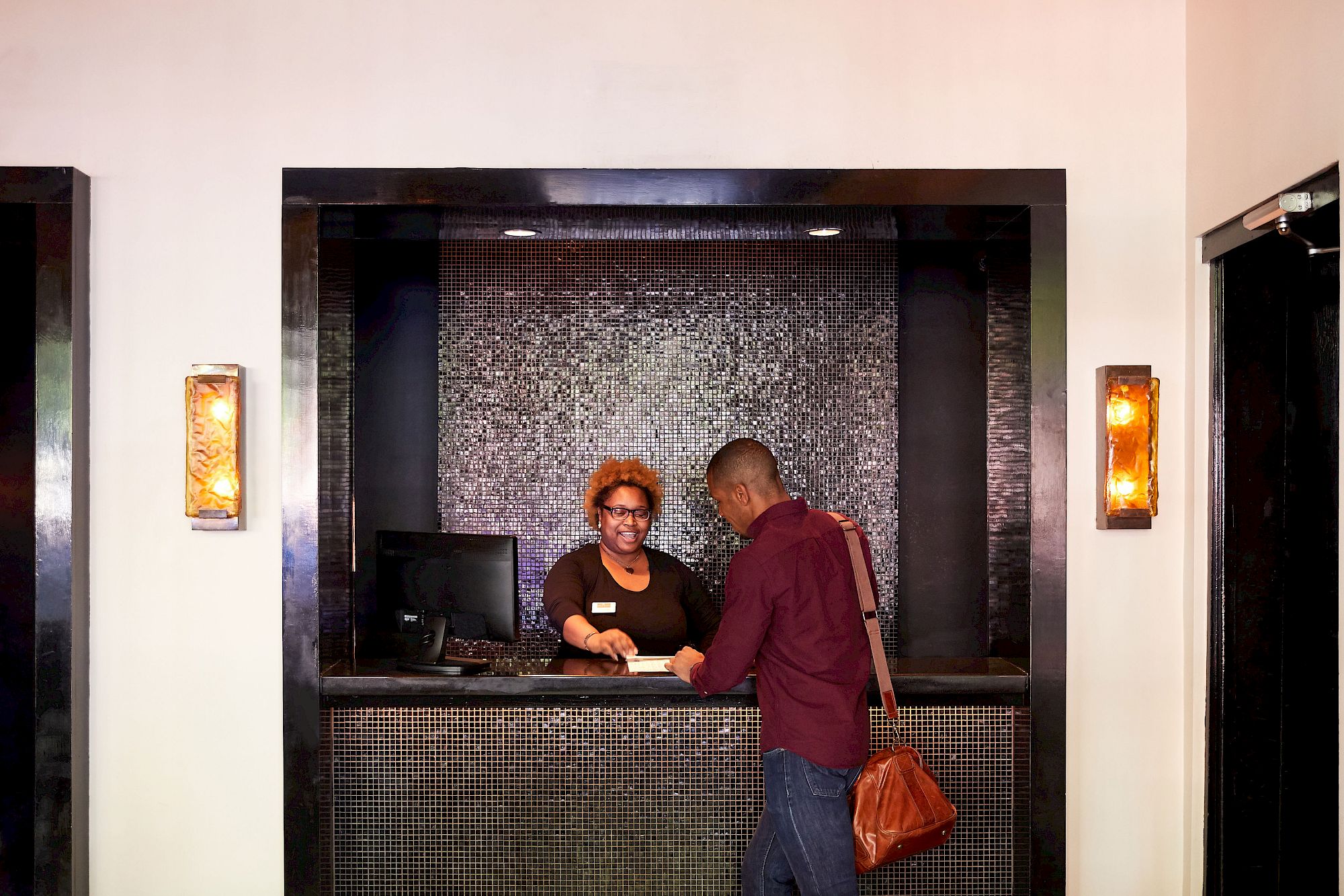 A man is interacting with a receptionist at a counter. The reception area has dark decor and soft lighting.