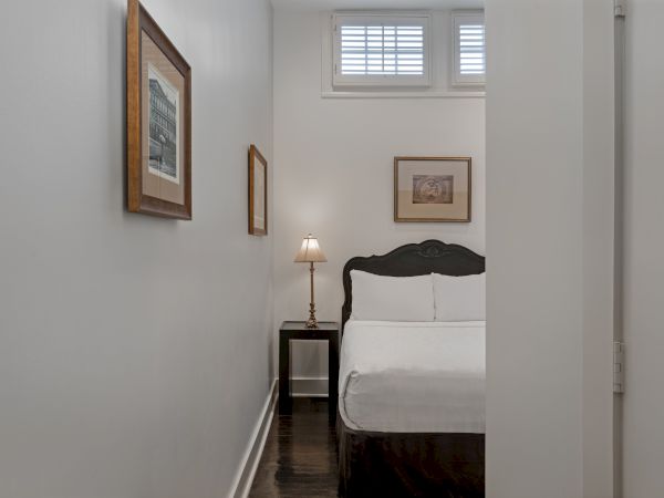 A small bedroom features a bed, a lamp on a nightstand, and framed pictures on the walls, with a window above the bed’s headboard.