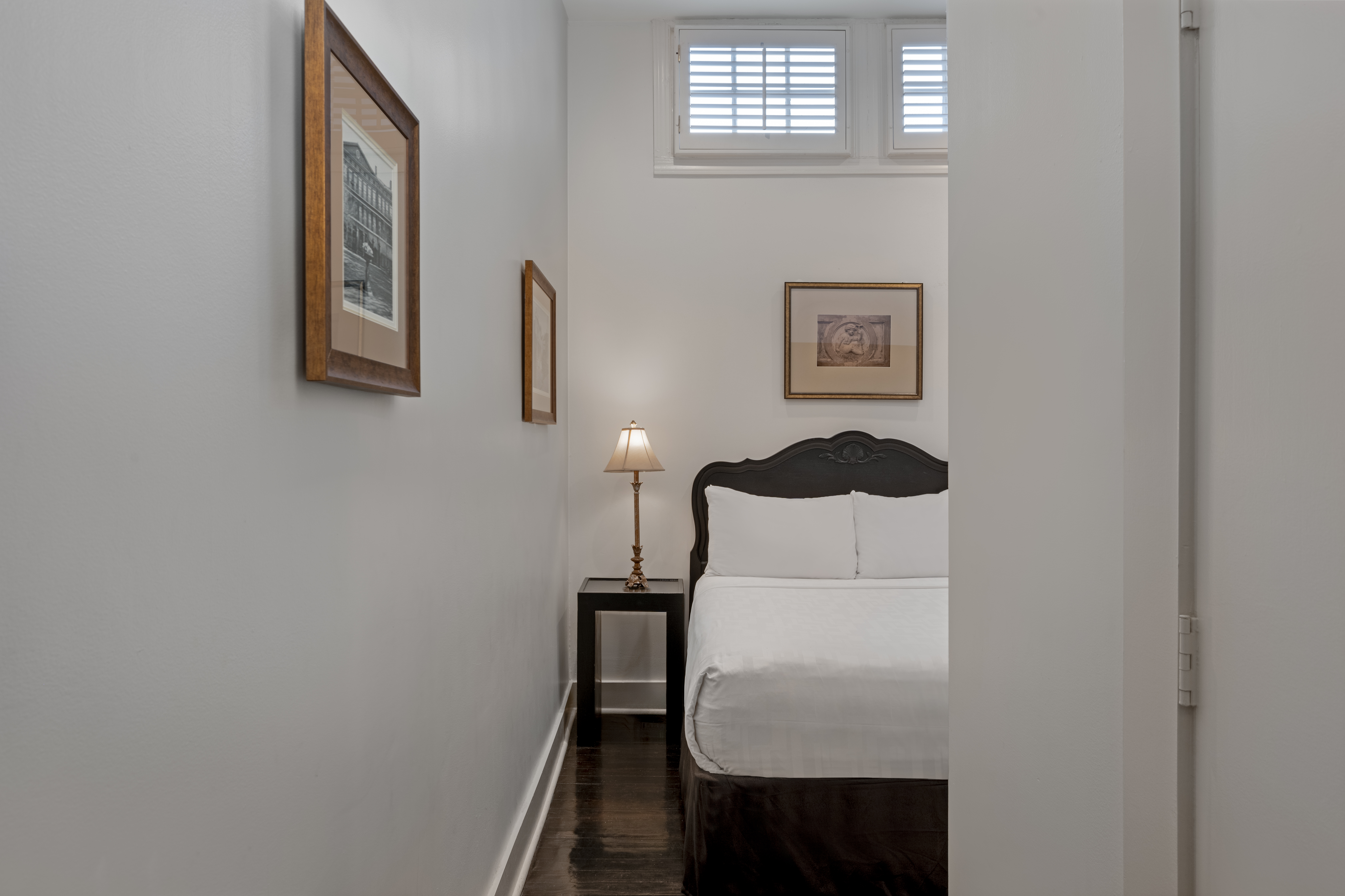 A neatly made bed with white linens and a dark wooden frame is centered in a small, white-walled room with framed art, a bedside table with a lamp, and a window above the bed.