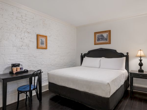 A cozy bedroom with a white brick wall, bed, side table, lamp, desk, chair, and framed pictures on the wall.