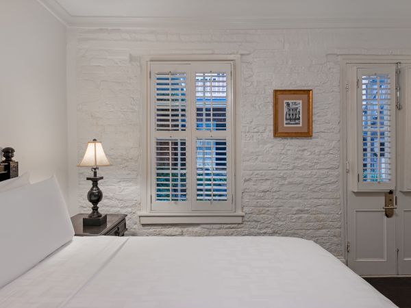 A cozy bedroom with a neatly made bed, a lamp on a nightstand, and windows with white shutters. A small framed picture decorates the wall.