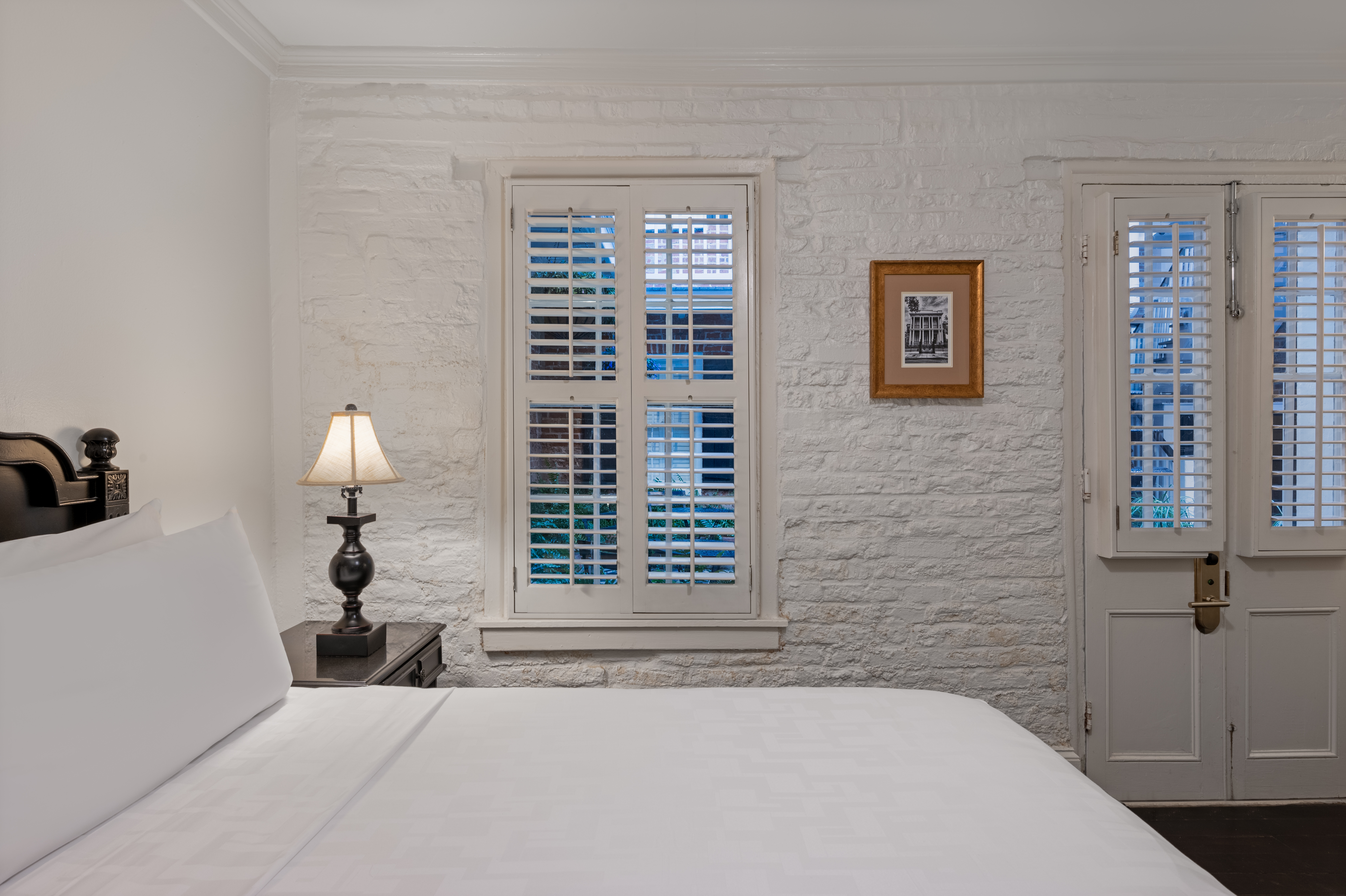 A white-painted brick-walled room with a bed, lamp, window, and door.