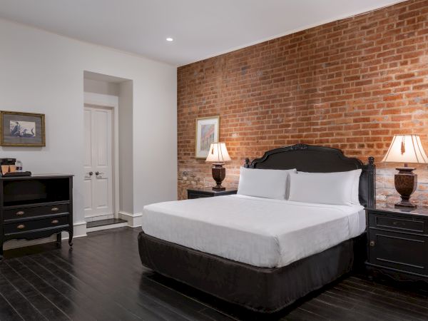 A bedroom with a brick wall, a large bed, two lamps on dark nightstands, and a dresser, creating a cozy and stylish atmosphere.