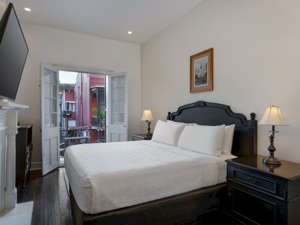 A cozy bedroom with a large bed, two lamps on side tables, a TV, and a window door opening to a balcony with a view outside.