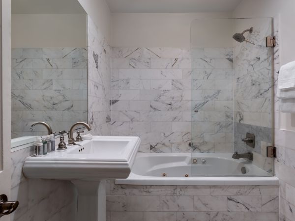 The image shows a bathroom with a white marble design, featuring a pedestal sink, bathtub with shower, glass partition, and mounted towels.