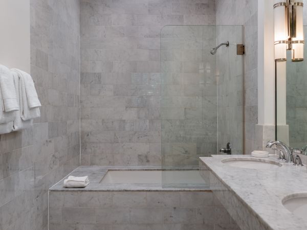 The image shows a modern bathroom with marble tiles, a glass shower, double sinks, and towels neatly arranged on a rack.