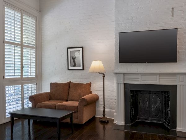 A cozy living room with a brown sofa, a black coffee table, a floor lamp, a wall-mounted TV above a fireplace, and a framed picture.