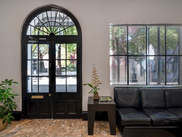 A cozy room with a black door, large window, plants, a black couch, and a small table, with natural light coming in.