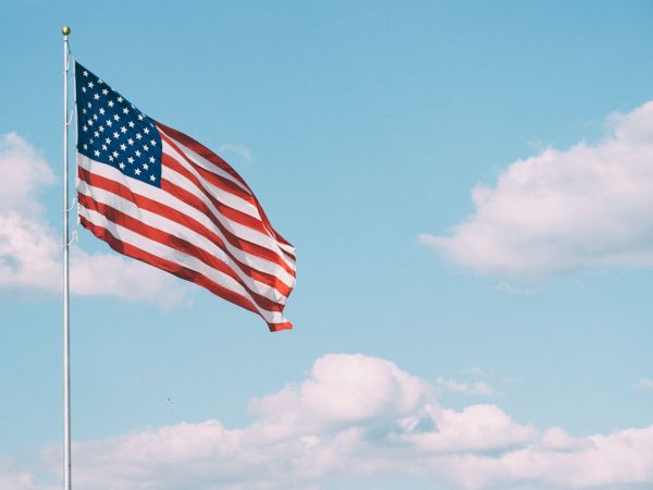 A U.S. flag waves against a clear blue sky with a few clouds visible in the background.