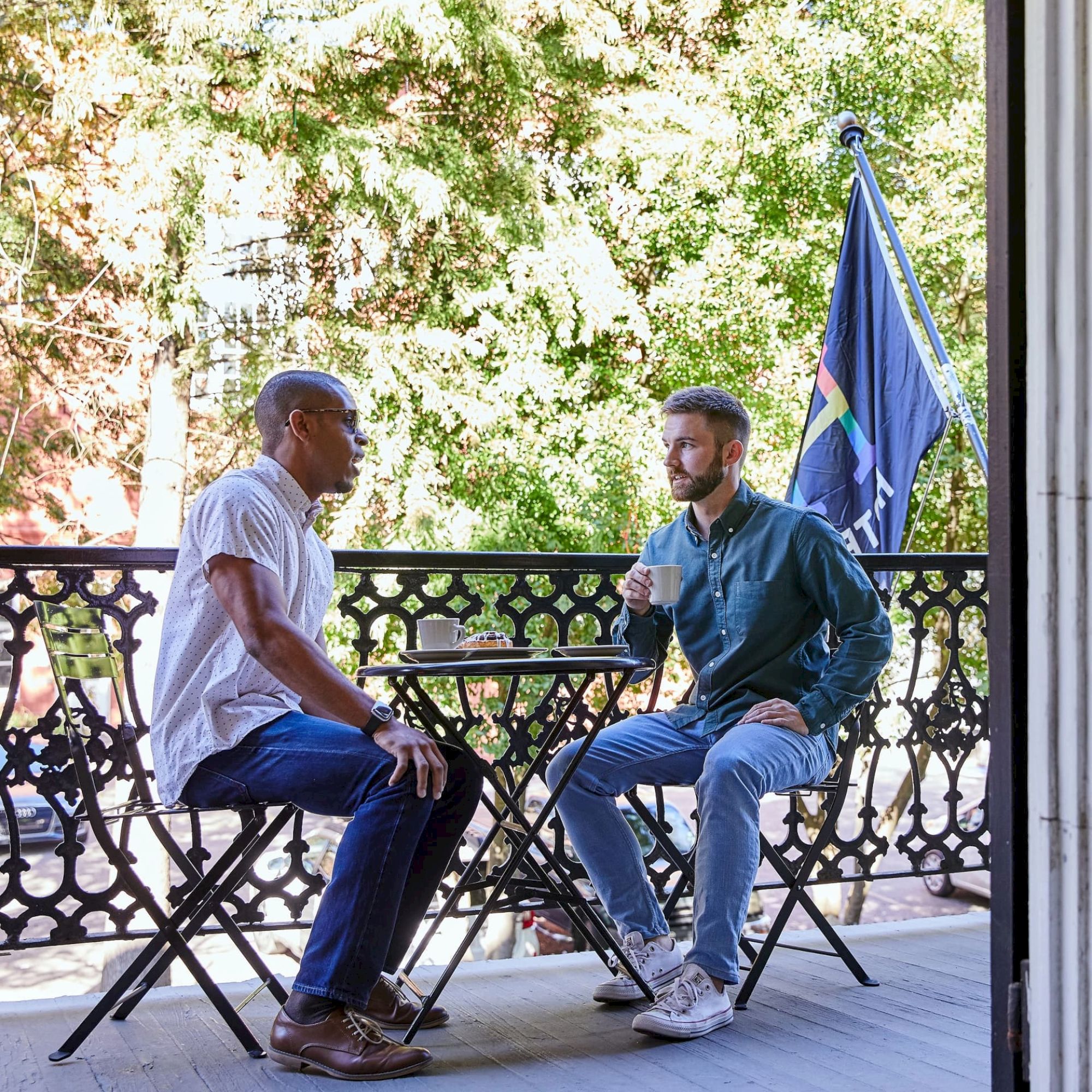 Two people sit on a balcony with an ornate railing, having a conversation over drinks. There's a flag in the background.