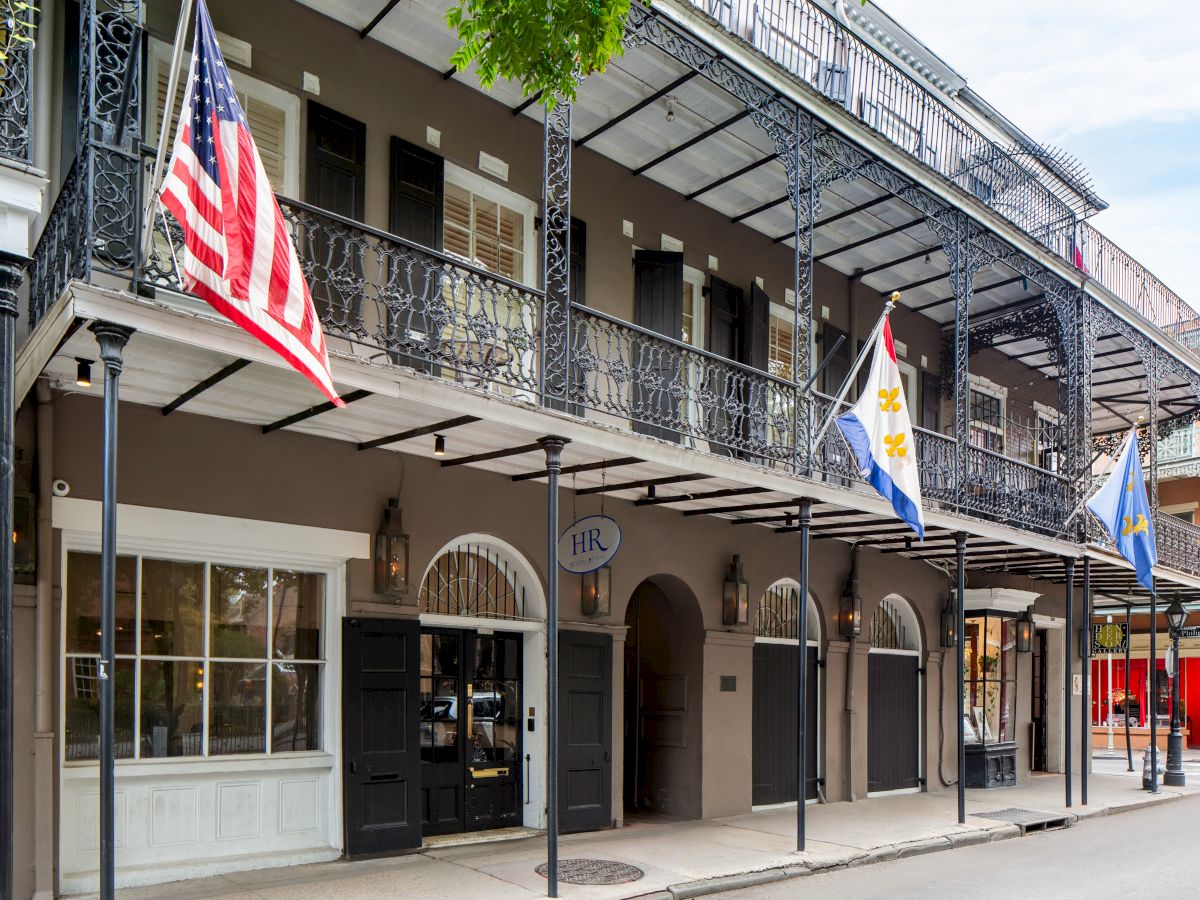 A historic building with wrought iron balconies, displaying three flags, stands on a quiet street, exuding Southern architectural charm.