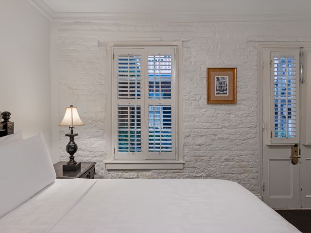 A white brick-walled room with a double bed, nightstand, lamp, and shuttered window.