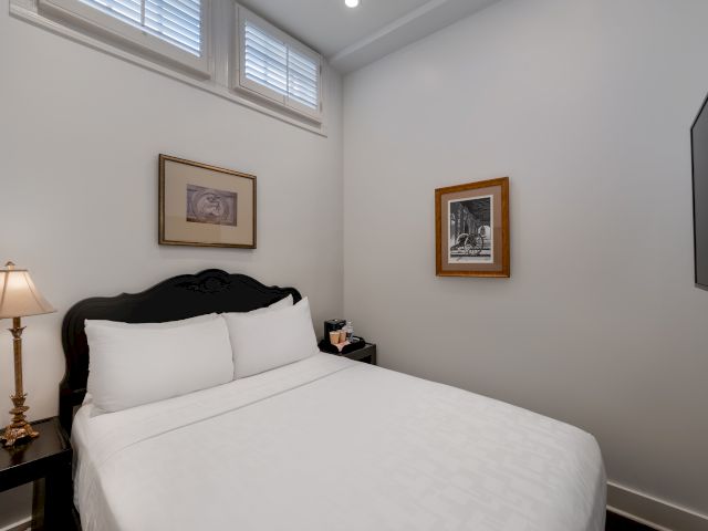 A neatly made bed with white linens and a dark wooden headboard in a small, white-painted room with two framed pictures on the walls and a window above the bed.