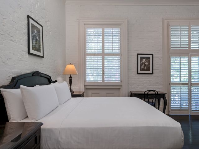 A neatly made bed with white linens and pillows in a room with white brick walls and dark wood furniture.