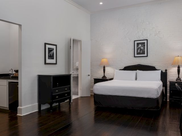 A wide view of a hotel room with a bed, nightstands, a dresser, and a doorway leading to a bathroom and kitchenette.