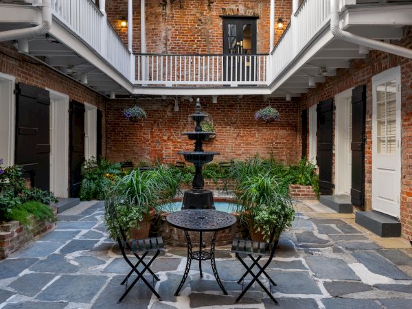 A courtyard with a central fountain, surrounded by brick walls, lush plants, and a small table with chairs on a stone floor.