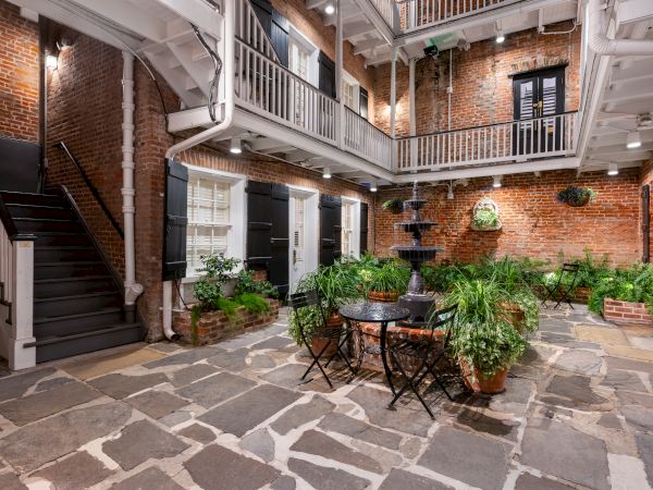 A brick-walled courtyard with stone floor, a small round table and chairs, potted plants, and a stairwell leading to an upper balcony.