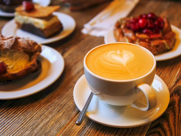 A cup of latte with latte art on a wooden table, surrounded by various pastries on plates in the background.