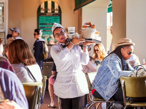 A waiter carries a tray with food and drinks through a busy cafe with seated patrons, capturing a lively atmosphere.